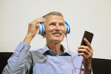 elderly man listen music with headphone. Portrait of a mature smiling man listening music on the black sofa at homeの写真素材