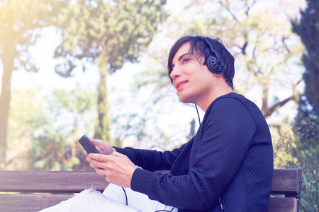 Joyful attractive guy listening to music on headphones enjoying in the park. Happy man listening to music from a smart phone with a against the bright sun. Toningの写真素材