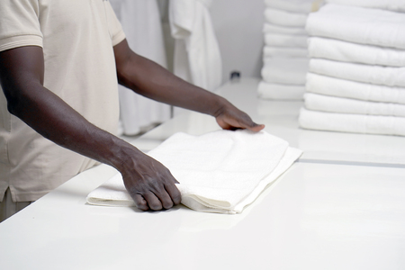 Hands of an african male laundry hotel worker folds a clean white towel. Hotel staff workers. Hotel linen cleaning services.の写真素材