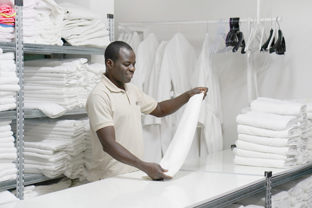 An African male hotel worker folds a clean white towel. Hotel staff workers. Hotel linen cleaning services.の写真素材