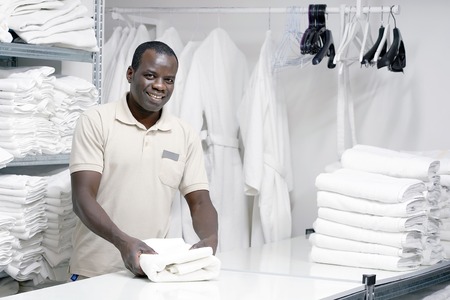 An African male hotel worker folds a clean white towel. Hotel staff workers. Hotel linen cleaning services.の写真素材