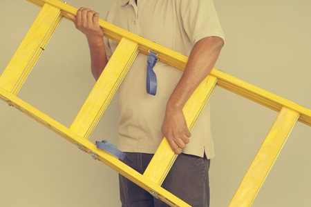 Electrician holds a yellow wooden ladder. The builder bears a wooden step-ladder on a gray background. Factory worker with a yellow wooden staircase. Toningの写真素材