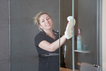 The maid in the bathroom wipes the glass of the shower room in the hotel room. Cleaning. Woman cleans the glass in the bathroomの写真素材
