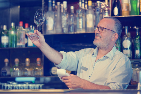 Elderly bartender cleans the glass. A handsome bartender polishes a glass of wine glasses. The concept of service. Toningの写真素材