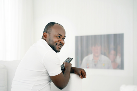Handsome young African man holding a remote control and laughing while sitting on the sofa at home....