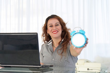 Red-haired woman holds an alarm clock in her hand in the workplace. female office worker holds an...