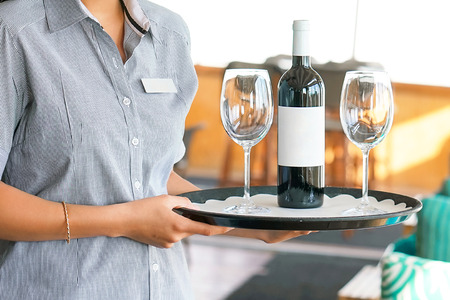 Young waitress carries a bottle of red wine with wine glasses on a tray to the customer of the hotel...
