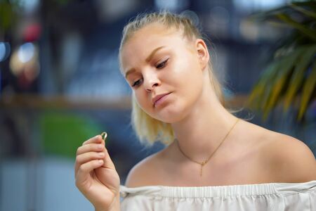 Young blonde girl holding wedding engagement ring in hands, engaged girl doubts about marriage proposal, abandoned wife depressed after getting divorced, help to overcome breaking up, starting new life.の写真素材