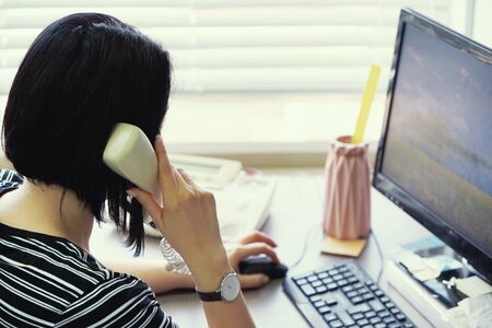 Close-up of office worker secretary answering phone calls and talking with customers, she is sitting at her desk working.の写真素材