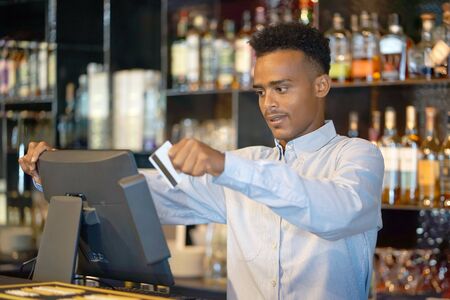 African bartender registration of a new order by a cash register. A restaurant worker registrating new order by cash-register. The barman pays the order with a credit card. Service concept.の写真素材