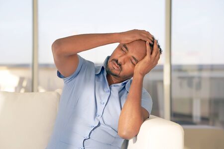 African Man suffering from headache migraine pain at home on sofa. Male holds head with hand. Concept of health.の写真素材