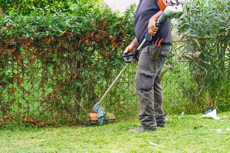 Professional gardener using an edge trimmer in the city parck. Elderly man worker mowing lawn with grass trimmer outdoors on sunny day.                              の写真素材