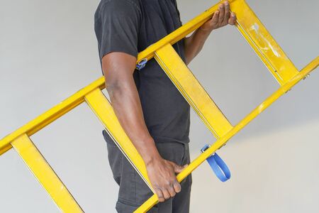 African electrician holds a yellow wooden ladder. The builder bears a wooden step-ladder on a gray background. Factory worker with a yellow wooden staircase.の写真素材
