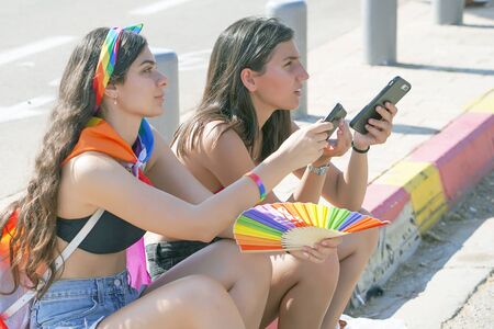 Two young lesbian girls are sitting on the road. Lesbians in the Gay Pride parade. Parade of tolerance. Rainbow flags at Gay pride parade. The annual parade LGBT. 14 June 2019. Tel Aviv. Israel.のeditorial素材