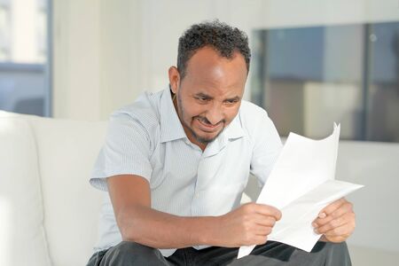 An agitated afro man without joy reads negative letter. African male reads negative news in a letter at home on the couch.の写真素材