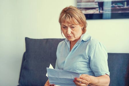 Ederly woman reads negative news in a letter at home on the couch. An elderly woman received a notice of the loss of a child, her husband.の写真素材