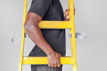 African electrician hand holds a yellow wooden ladder. Factory african worker with a yellow wooden staircase.The builder bears a wooden step-ladder on a gray background.                                           の写真素材
