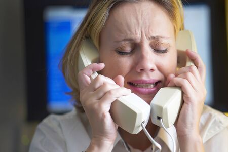 Face of office woman in stress with a lot of phone calls at same time. Unhappy woman holds two handsets of the phone. Busy office worker female working and answering a lot of calls at the same time.の写真素材