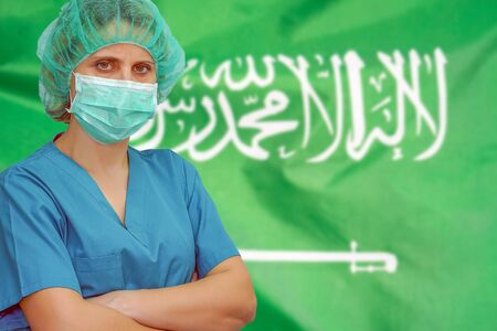 Female surgeon in mask and hat looks at the camera on the background of the Saudi Arabia flag. Health care and medical concept. Surgery concept and fight the virus in Saudi Arabia.の写真素材