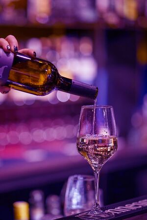 Hand a bartender woman pours white wine into a glass from a bottle. Night time. Toning