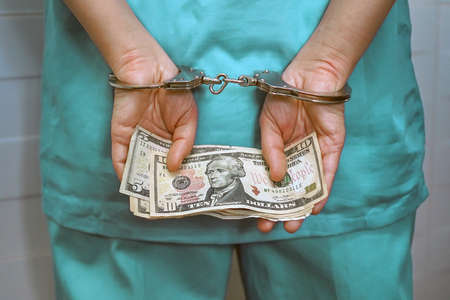 Close-up crime in medicine, surgeon or doctor in green uniform with handcuffed hands holding a bribe. Criminal medic woman hands locked in handcuffs. Back viewの写真素材