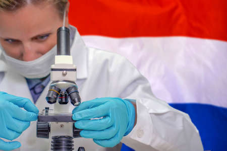 Female biochemist looking through a microscope against Netherlands flag background. Medical technology and pharmaceutical research and development of science concept in Dutchの写真素材