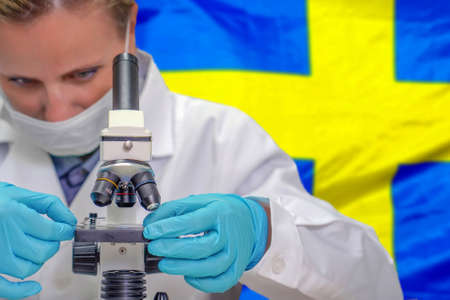 Female biochemist looking through a microscope against Sweden flag background. Medical technology and pharmaceutical research and development of science concept in Swedenの写真素材