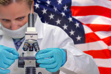 Female biochemist looking through a microscope against US flag background. Medical technology and pharmaceutical research and development of science concept in USAの写真素材