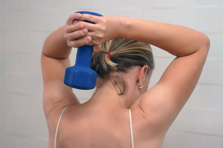 Woman holding a dumbbell from the head. Active senior female exercising with dumbbells. Rear view of...
