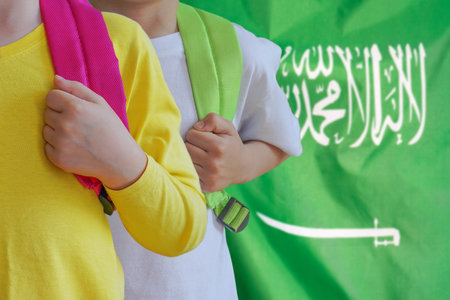 Two children with satchels background of Saudi Arabia flag. Concept of upbringing and educating children in Saudi Arabiaの写真素材