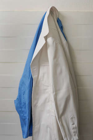 White and blue smock on wooden hanger on ceramics wall background. Closeup of doctor's scrubs and lab coat on hangersの写真素材