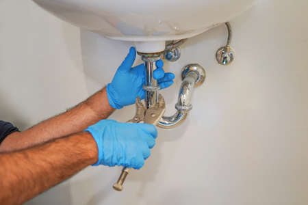Plumber with blue gloves repairing sink siphon in kitchen. Plumber repairs and maintains chrome siphon under the washbasin. Refurbishment in the apartment. Elimination of leakage in the sinkの写真素材