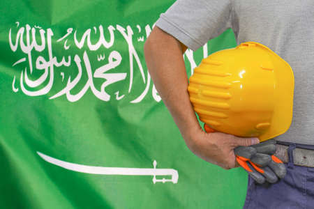 Close-up of hard hat holding by construction worker on Saudi Arabia flag background. Hand of worker with yellow hard hat and gloves. Concept of Industry, construction and workers in Saudi Arabiaの写真素材
