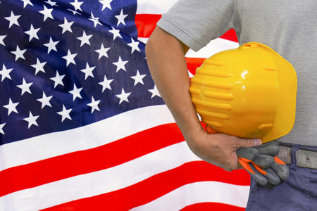 Close-up of hard hat holding by construction worker on US flag background. Hand of worker with yellow hard hat and gloves. Concept of Industry, construction and industrial workers in USAの写真素材
