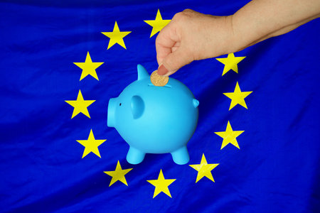 Hand of elderly woman putting coin into piggy bank on EU flag background. Hand putting coin to piggy bank. Retirement savings, piggy bank. Concept saving money, investment and retirement fund in EUの写真素材