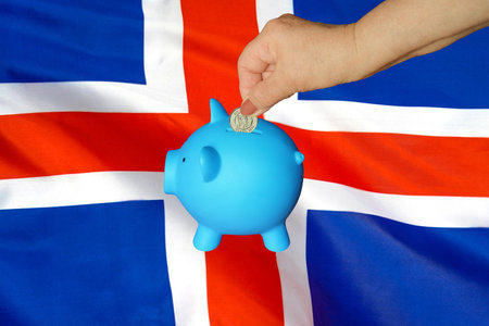 Hand of elderly woman putting coin into piggy bank on Iceland flag background. Hand putting coin to piggy bank. Retirement savings, piggy bank. Concept saving money and retirement fund in Icelandの写真素材
