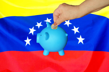 Hand of elderly woman putting coin into piggy bank on Venezuela flag background. Hand putting coin to piggy bank. Retirement savings, piggy bank. Concept saving money and retirement fund in Venezuelaの写真素材