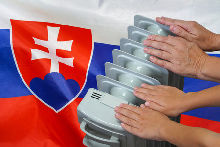 Hands of mother and son near a hot home radiator are trying to warm themselves on Slovakia flag background. Hands of woman and teen near cold home radiator. Cold season and gas crisis in Slovakiaの写真素材