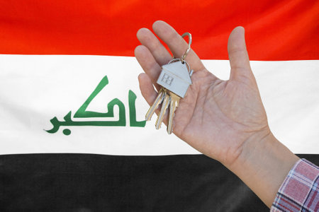 Hand shows the keys to the apartment on background of flag Iraq. Keys to the apartment in a female hand on background of flag Iraq. Realty concept and home buying or new apartment in Iraqの写真素材