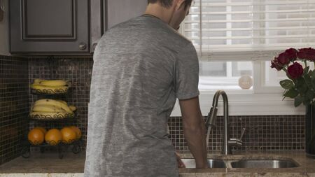 young attractive man in a gray t-shirt stands with his back to the camera and washes dishes or hands...