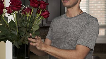 Young handsome man with a little stubble in a gray T-shirt straightens fixing and sniffs a beautiful bouquet of red roses that stands in a blue vase on a granite table in a fashionable kitchenの写真素材