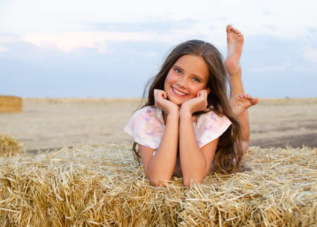 Adorable happy smiling ittle girl child lying on a hay rolls in a wheat field at sunsetの写真素材