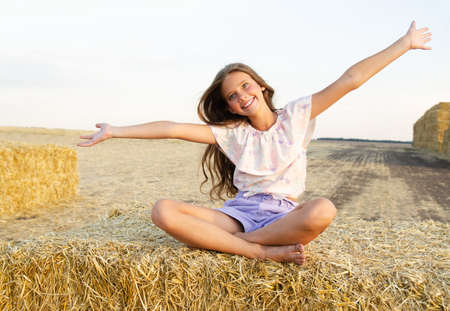Adorable happy smiling ittle girl child sitting on a hay rolls in a wheat field at sunsetの写真素材