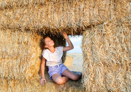 Adorable happy smiling ittle girl child sitting on a hay rolls in a wheat field at sunsetの写真素材