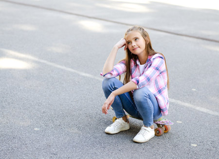 Smiling cute little girl child sitting with a skateboard and thinking. Preteen with penny board outdoors in summer dayの写真素材