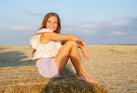 Adorable happy smiling ittle girl child sitting on a hay rolls in a wheat field at sunsetの写真素材