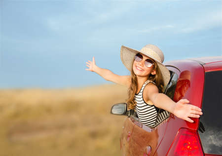 Happy smiling little girl child goes to summer travel trip in the red car. Preteen in hat and sunglasses enjoying road trip.の写真素材