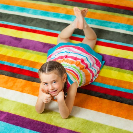 Happy child lying on the color carpet background and showing thumbs up. Little girl smiling and looking at cameraの写真素材