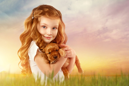 Beautiful red-haired little girl with red puppy in field at sunset. Fashion, style kid and pet friendshipの写真素材