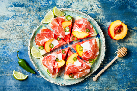 Fresh summer watermelon pizza with feta cheese, peach, prosciutto, jalapeno and honey drizzle on blue backgroundの写真素材